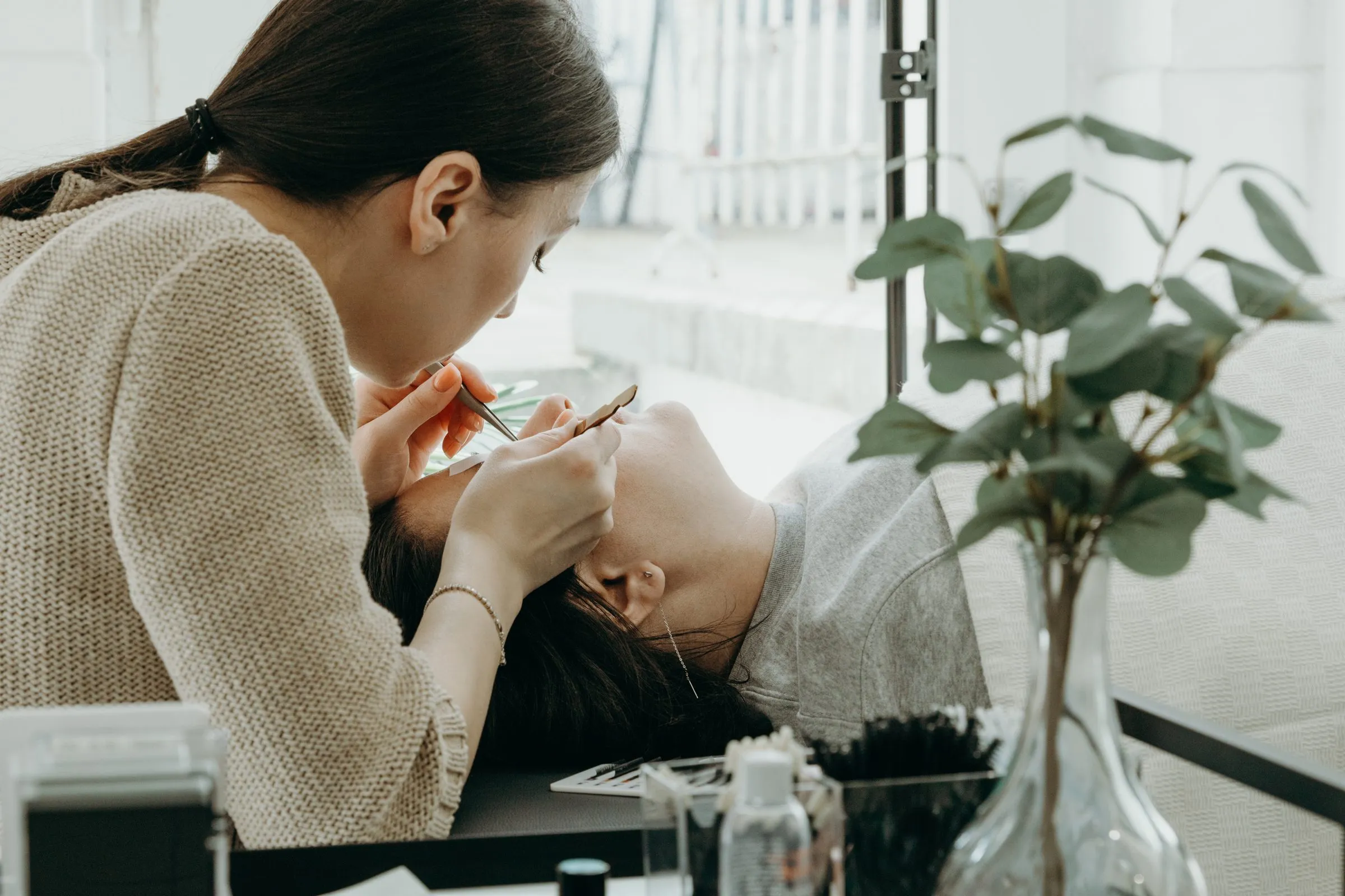Wimpernstylistin arbeitet am Fenster mit zwei Pinzetten an den Wimpern einer liegenden Kundin, im Vordergrund ein Eukalyptuszweig